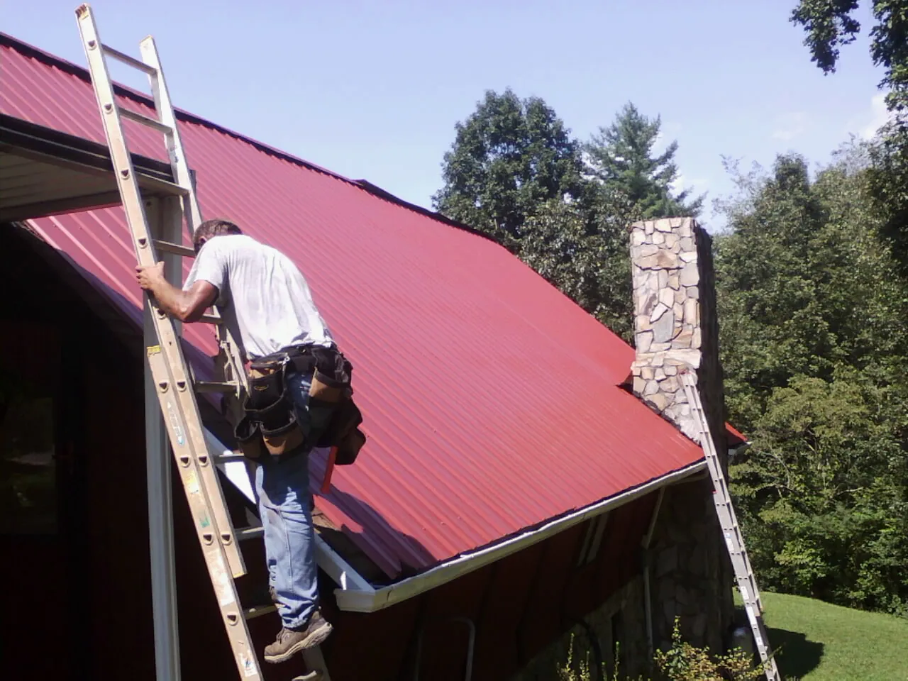 Roofer on a ladder beside a red metal roof and stone chimney — Soffit & Fascia Repair by Roofing Brothers in Los Alamitos